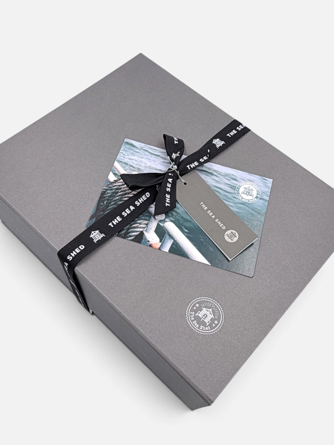 Closed grey Sea Shed gift box with black branded ribbon, gift tag and coastal postcard